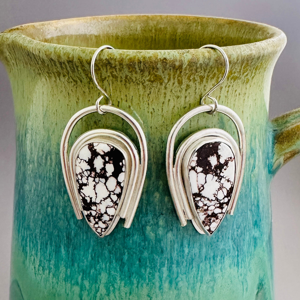 A pair of silver dangle earrings with reverse teardrop-shaped white and brown marbled stone centers, hanging in front of a green and teal ceramic pitcher.
