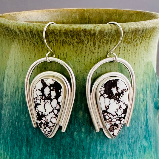 A pair of silver dangle earrings with teardrop-shaped Wild Horse Magnesite pendants, hanging in front of a green and teal ceramic vase.