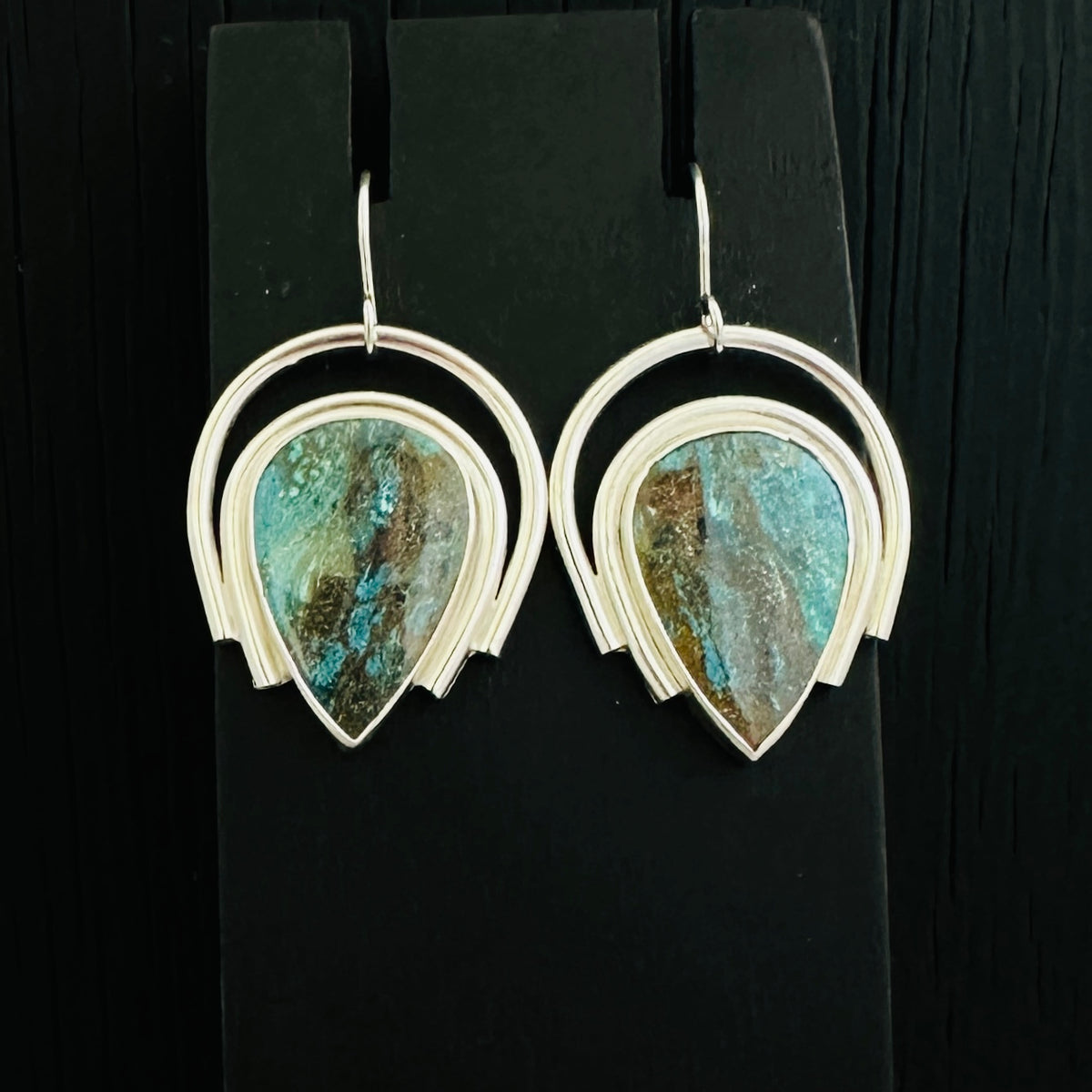 A pair of silver hoop dangle earrings with teardrop-shaped turquoise and brown stone pendants, displayed on a black stand against a dark wood background.